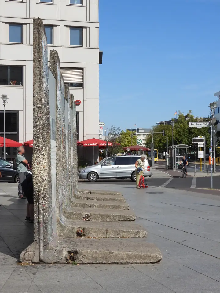 Four Berlin Wall elements seen edge-on on a city square, their broad concrete feet projecting to one side to form an L-shape.