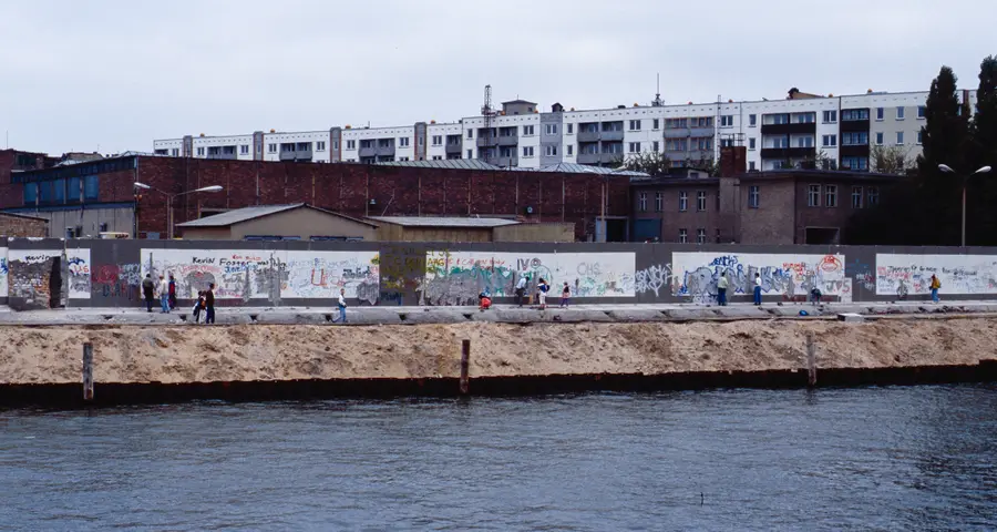 A long painted wall runs along a river embankment in front of brick industrial buildings and apartment blocks, with people walking along it.