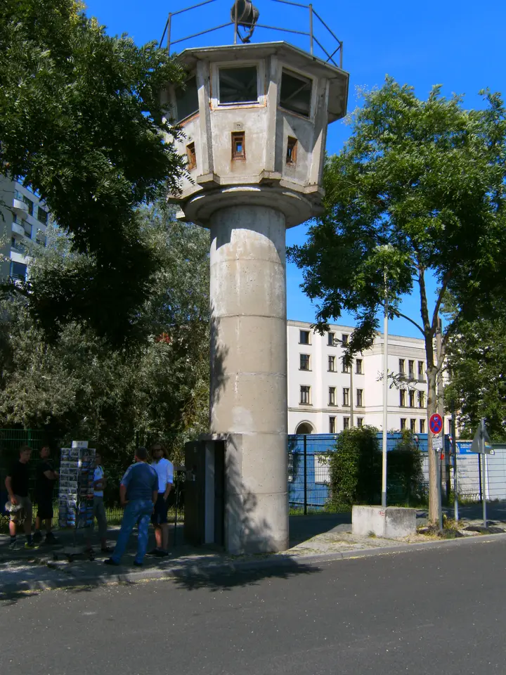 A slender concrete watchtower with a round shaft and a wider octagonal observation cabin with windows, standing among trees and modern office buildings.