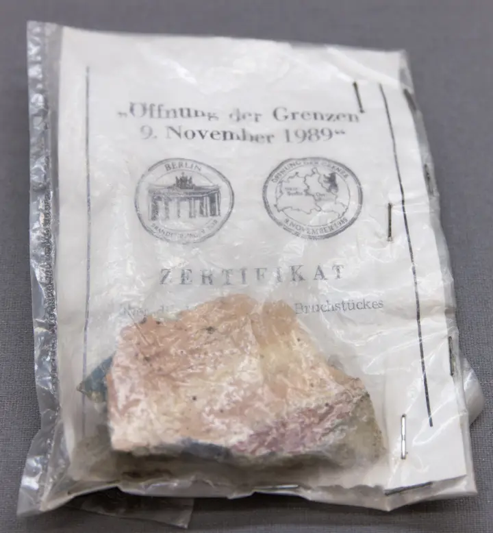 A small pinkish lump of concrete sealed in a stapled plastic bag with a printed card headed 'Öffnung der Grenzen 9. November 1989' and 'Zertifikat'.
