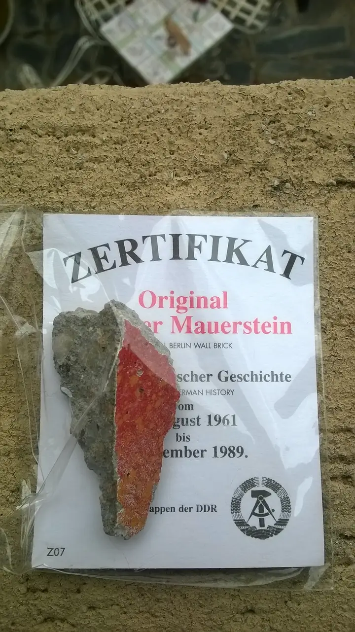 A small grey fragment of concrete with one face painted bright red, mounted on a card headed 'Zertifikat' and 'Original Berliner Mauerstein' with dates from August 1961 to November 1989.