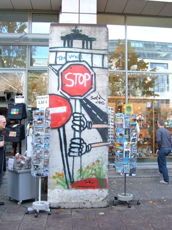 A single wall element painted with a stop sign, a hand and the Brandenburg Gate stands outside a souvenir shop between two racks of postcards.