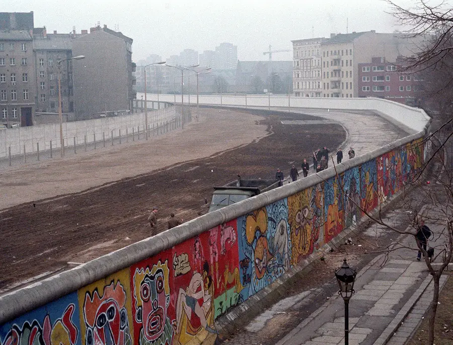 Seen from above, a curving stretch of the Wall covered in brightly painted faces and figures, with the raked death strip, a patrol track and a second, plain wall behind it and East Berlin apartment blocks beyond.