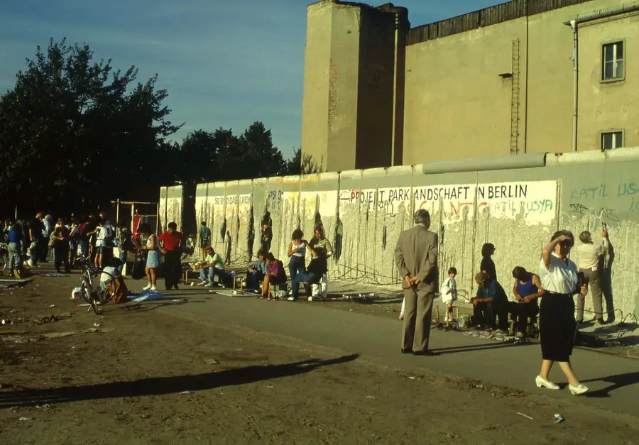A long wall stripped of most of its surface, with bent reinforcing bars hanging loose, while people sit and stand along it with hammers and small stalls in the evening sun.
