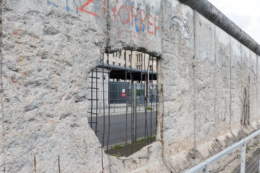 Close view of a surviving stretch of the Wall with a large hole broken through the concrete, exposed reinforcing bars crossing the gap and a fence and buildings visible through it.