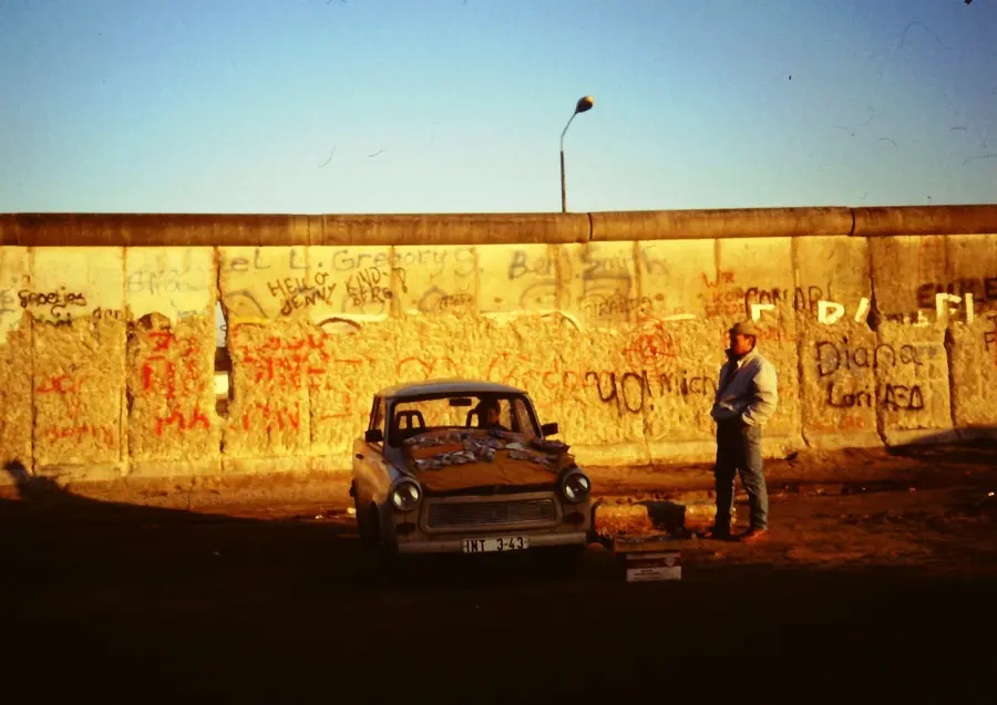 A man stands beside a Trabant parked in front of the graffitied Wall at sunset, with fragments of concrete laid out for sale on the car's bonnet.