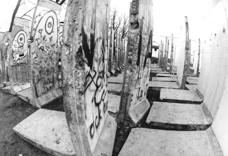 Black-and-white wide-angle view down rows of L-shaped wall segments standing on their feet on bare ground, several covered in graffiti.