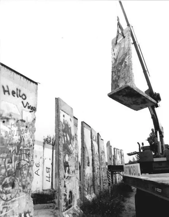 Black-and-white photograph of a crane lifting a graffitied L-shaped wall segment onto a lorry, with a row of numbered segments standing on the left.