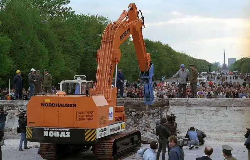An orange tracked excavator with a hydraulic hammer breaks up a thick concrete wall into rubble while crowds watch from the other side and border guards stand on top.