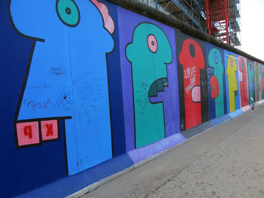 A row of large flat-coloured cartoon heads in profile, in blue, green, red and yellow, painted along a stretch of wall beside a pavement.