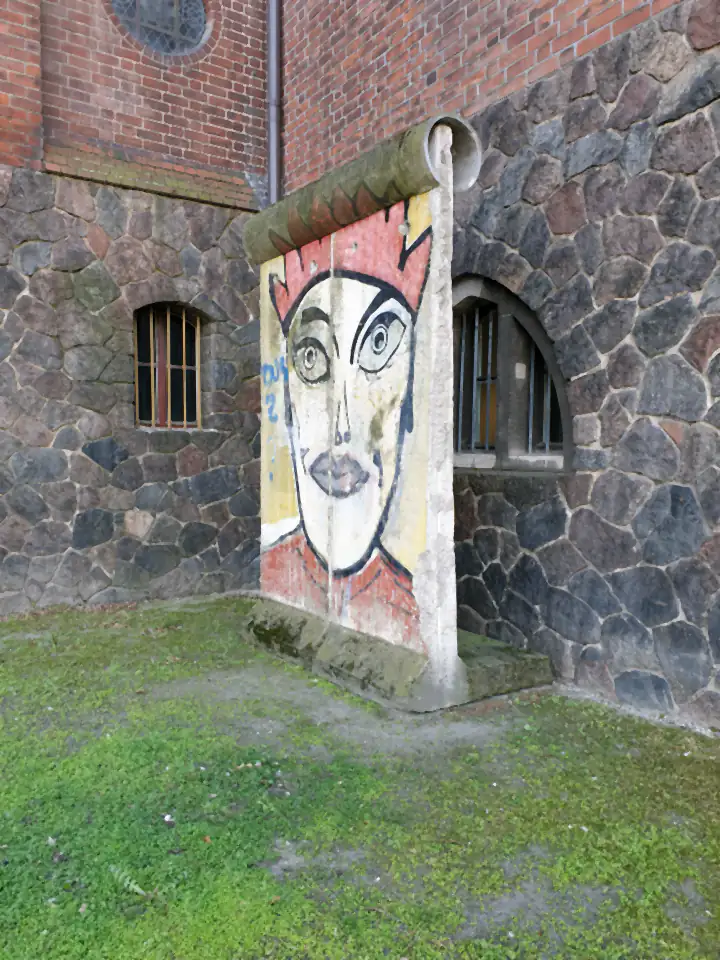 A single wall element standing against a brick and stone building, painted with a large stylised face wearing a red crown.