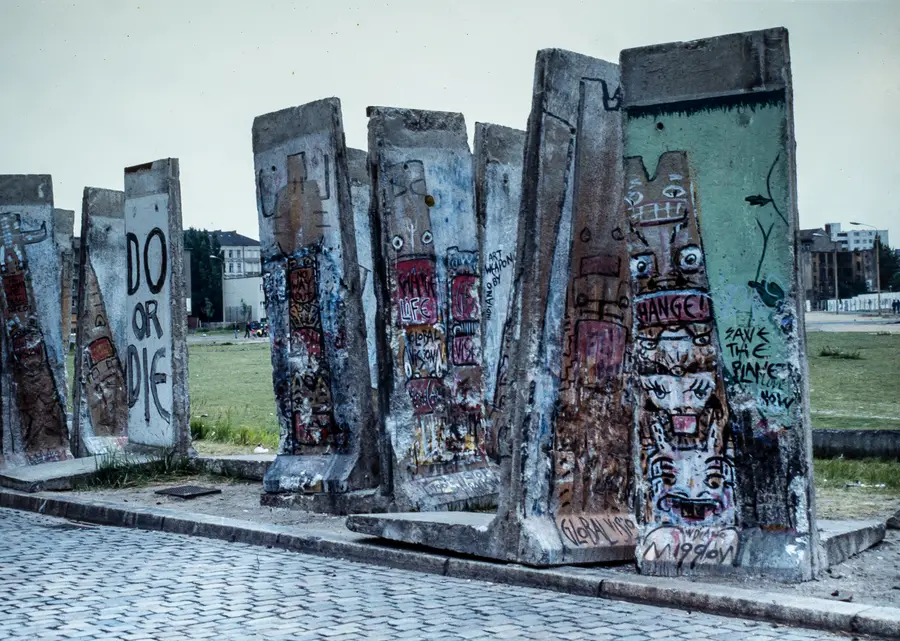 A cluster of dismantled wall elements standing upright at odd angles on a cobbled roadside, painted with faces and slogans including 'Change', 'Global Vision' and 'Save the planet now'.