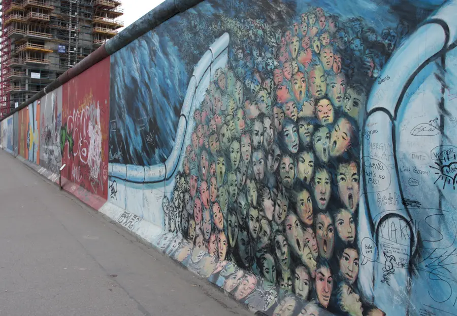A mural of a dense crowd of faces surging through a gap in a painted wall, set against a blue background, along a stretch of the East Side Gallery.