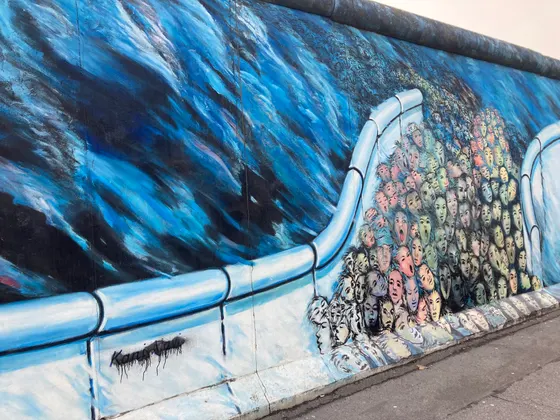 A crowd of faces pressing through a breach in the Wall, painted in blues and pale tones on the East Side Gallery.