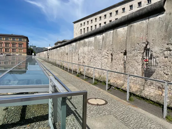 A long unpainted run of the Berlin Wall on Niederkirchnerstraße, its face chipped back to the reinforcing bars, beside the glass-roofed Topography of Terror trench, with the former Air Ministry building behind.