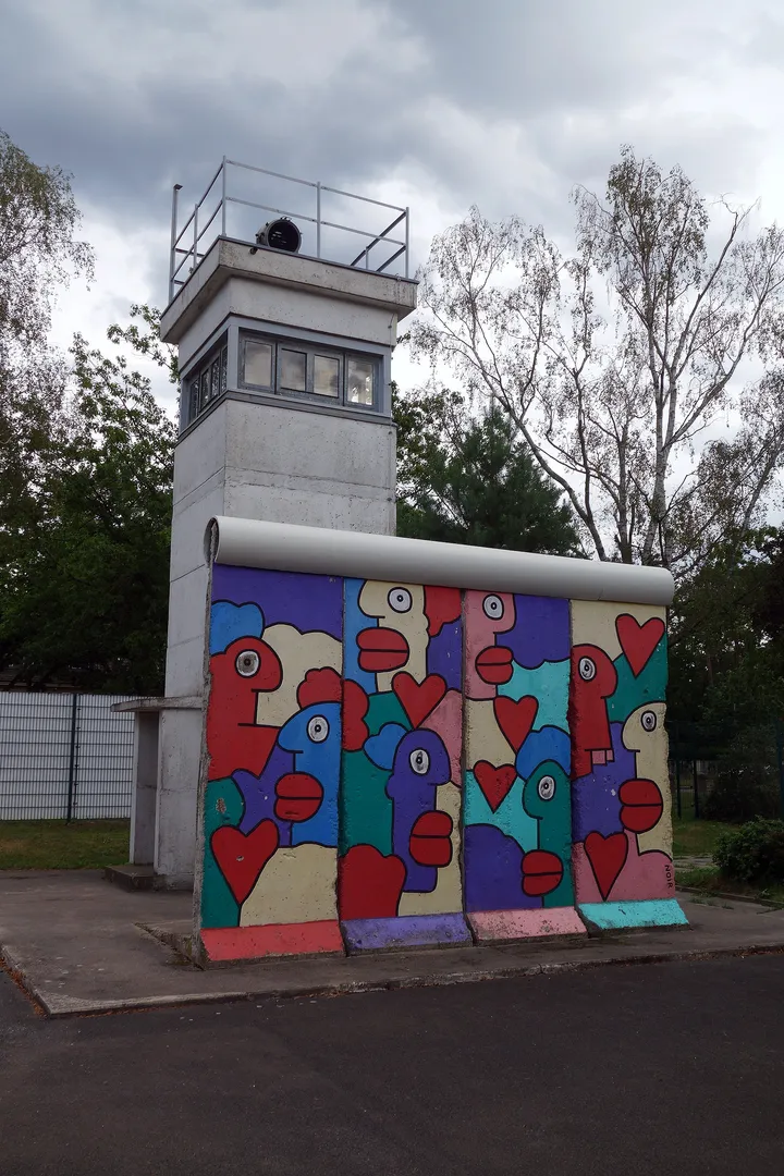 A section of the Berlin Wall standing beside a square concrete border watchtower on grass.