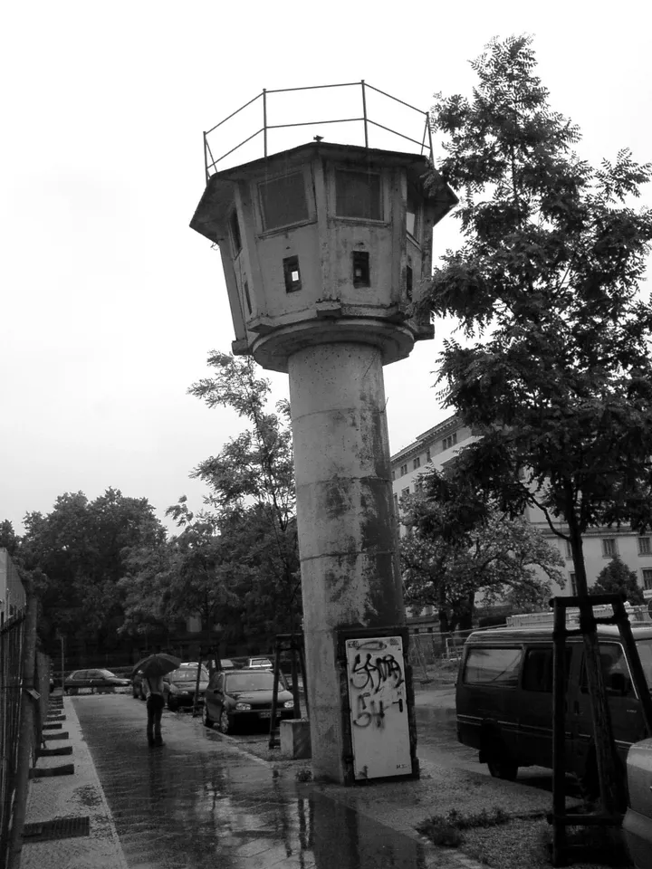 A slender round concrete tower with a glazed octagonal cabin on top, standing between modern buildings.