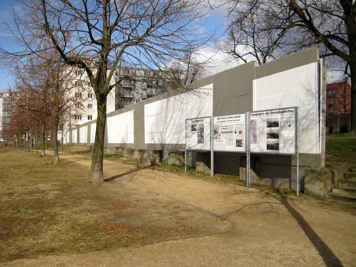 A white-painted concrete wall and a paved patrol strip running between trees in a cemetery.