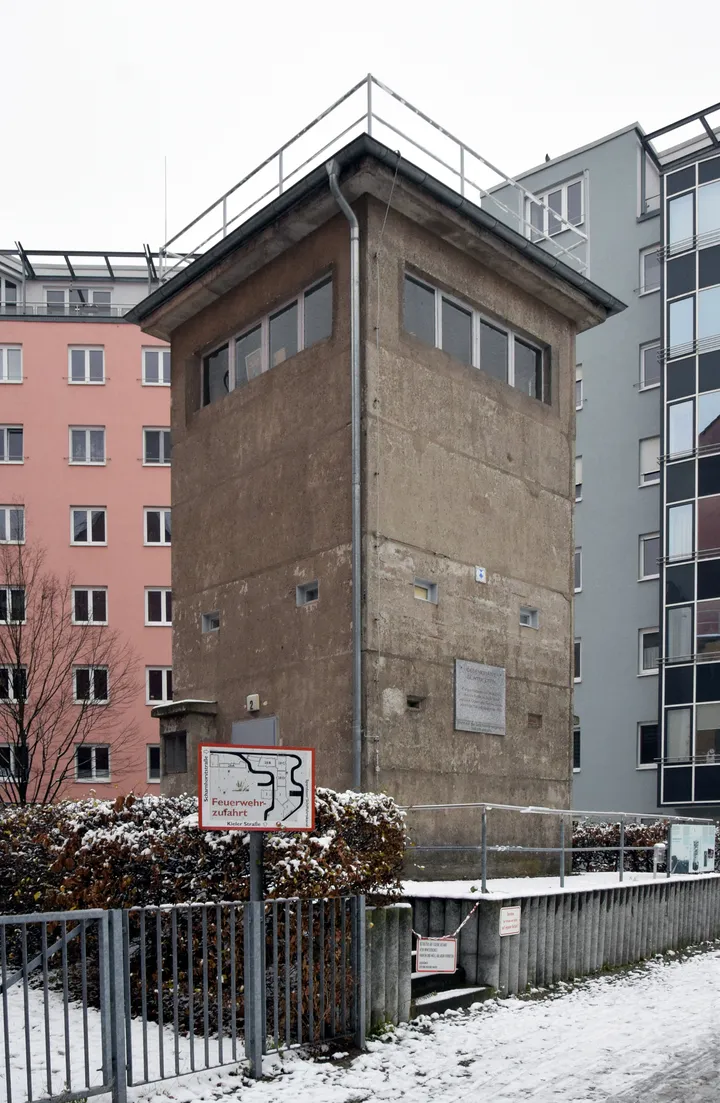 A square concrete border watchtower standing among trees beside a canal.
