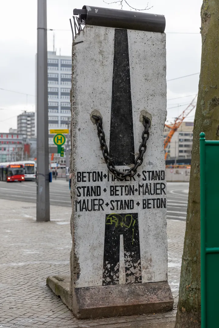 A painted Berlin Wall segment standing on a paved square outside a railway station.