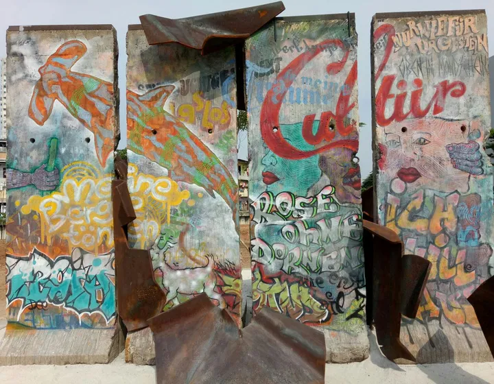 Painted Berlin Wall sections with cut metal figures climbing between them, in a park.