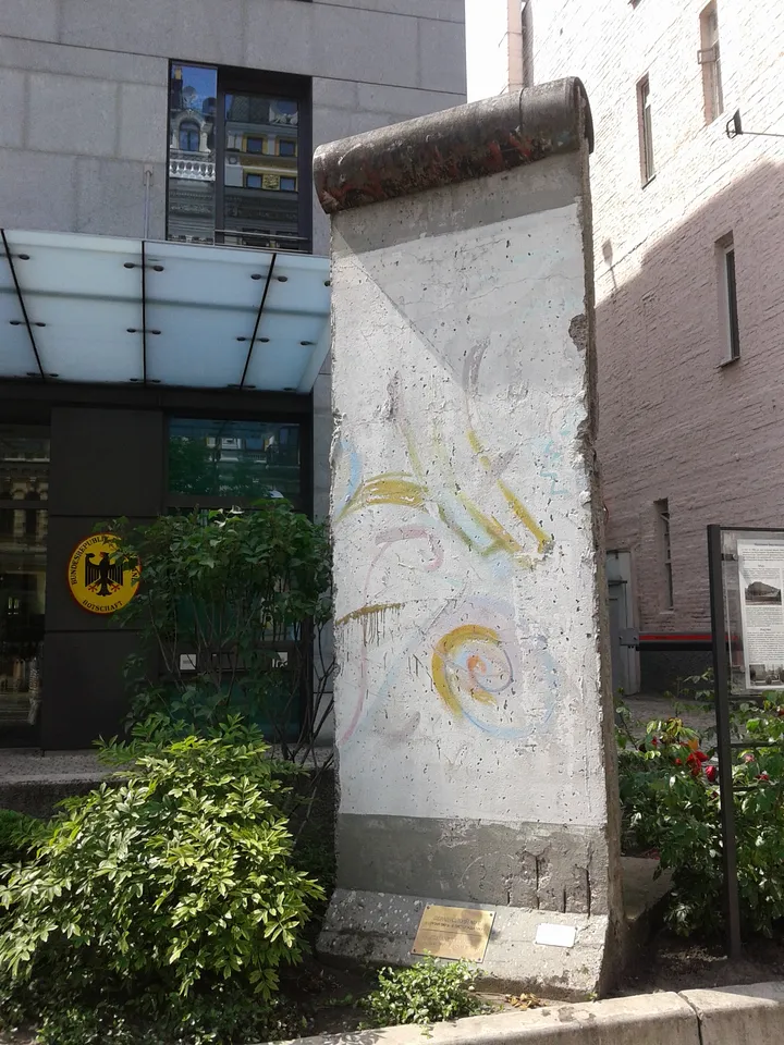 A painted segment of the Berlin Wall standing on a pavement beside railings.