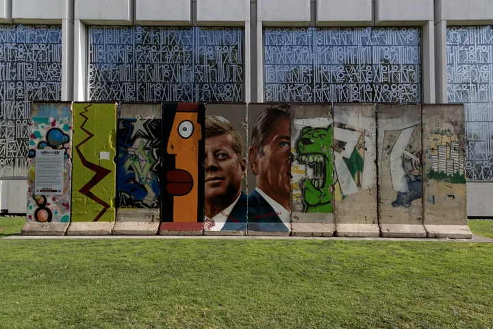 A long run of Berlin Wall segments painted with large faces, standing behind a street kerb.
