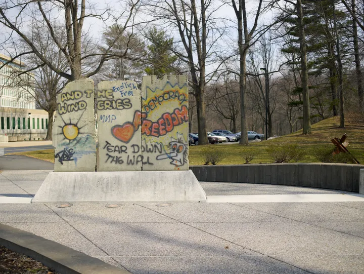 Three graffitied Berlin Wall sections on a low concrete plinth beside a path, with bare trees and a low building behind.