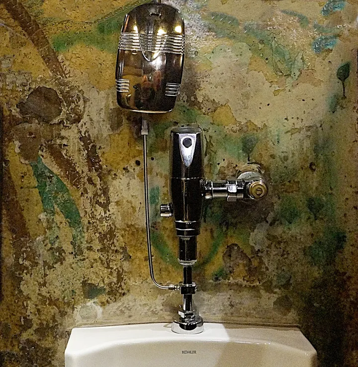 A graffitied Berlin Wall slab mounted behind glass in a tiled washroom, with urinals in front of it.