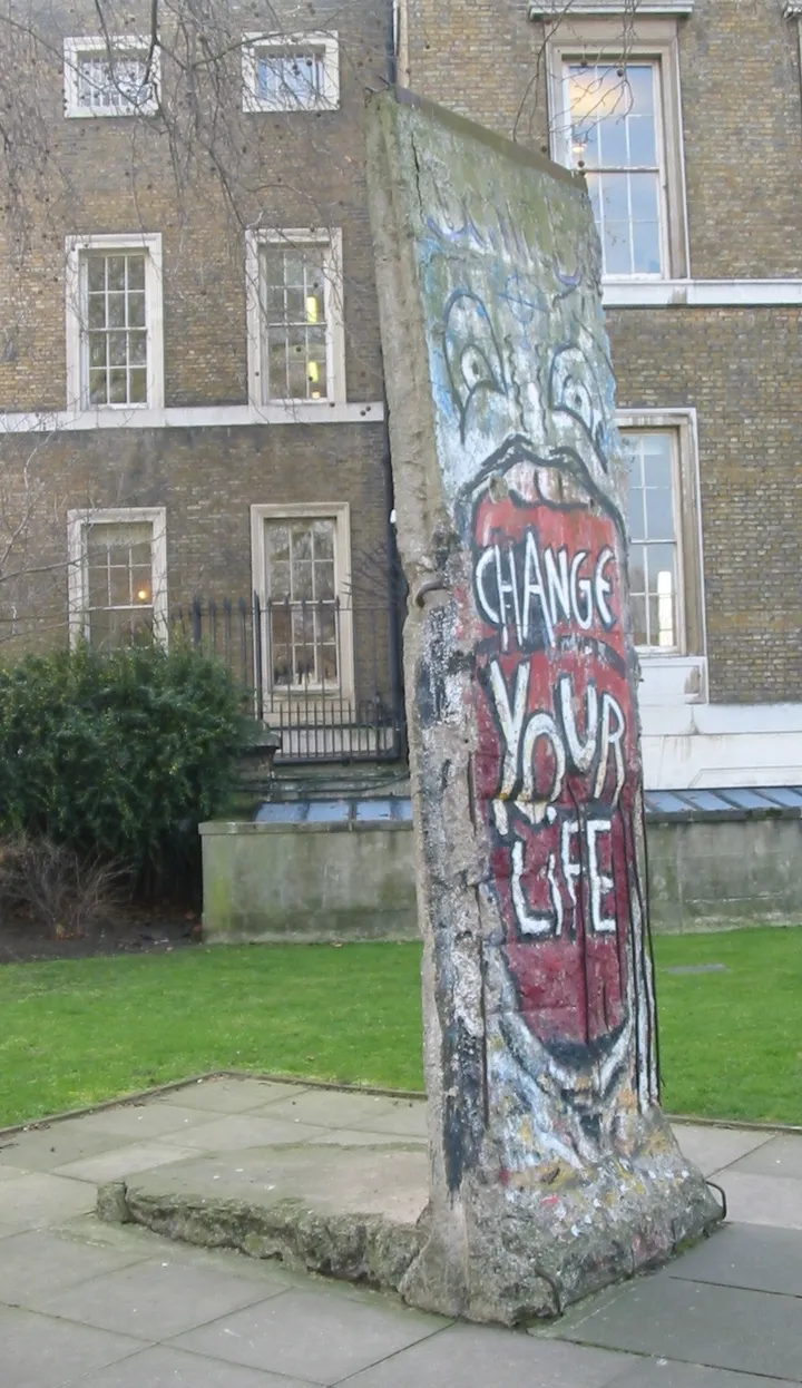 A Berlin Wall segment painted with an open red mouth reading CHANGE YOUR LIFE, standing in a park.
