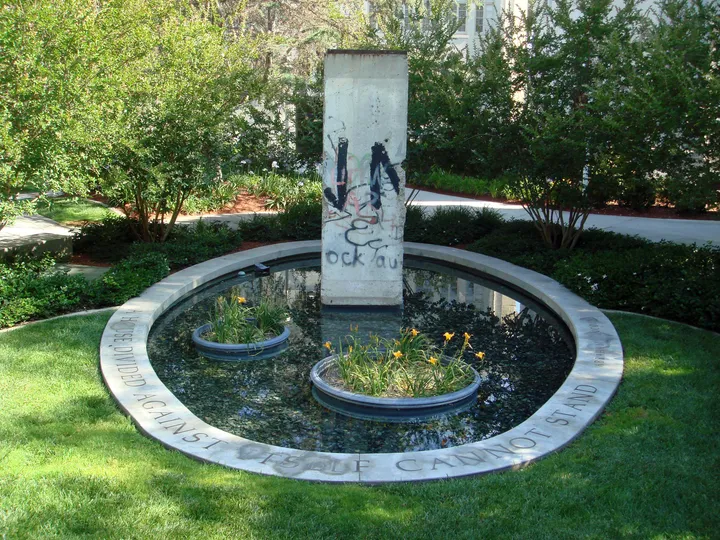 A Berlin Wall section standing in a shallow pond surrounded by paving.