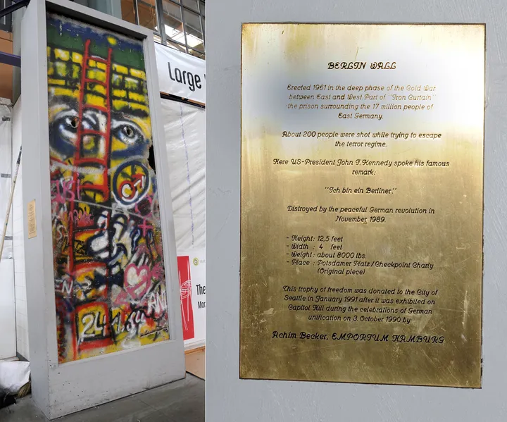 A painted Berlin Wall section with a commemorative plaque, standing indoors in a food hall.