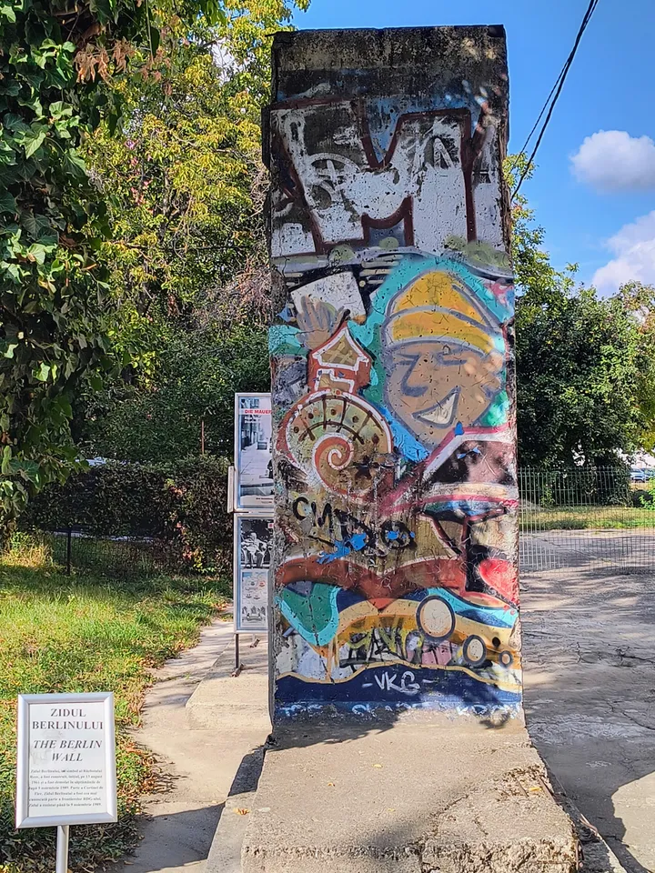 A tall concrete Berlin Wall segment covered in colourful graffiti, a smiling figure in a yellow cap above a spiral shell, standing on a concrete base beside a path with an information sign in the foreground.