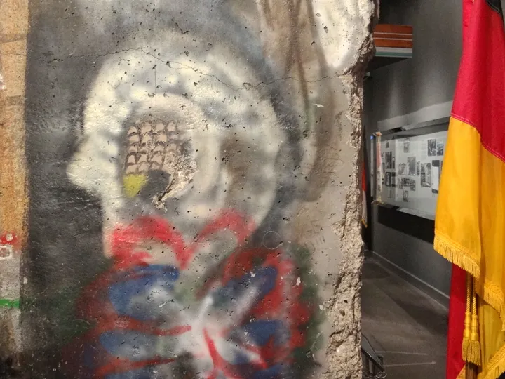 A painted section of the Berlin Wall displayed indoors beside a hanging German flag.